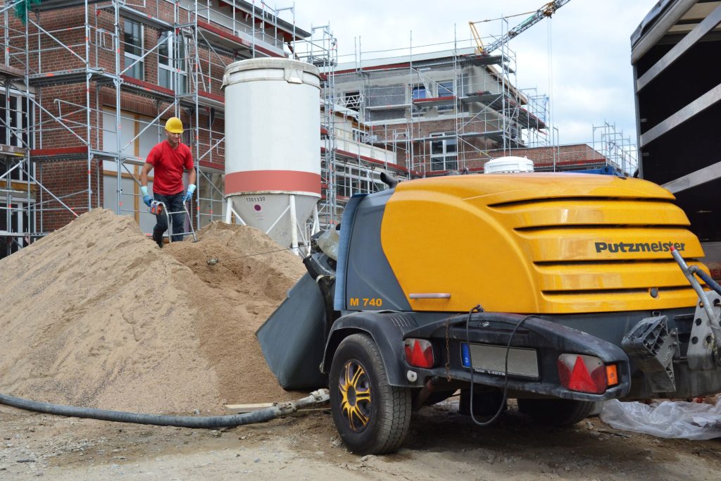 Gele Putzmeister M 740 mortelpomp op een bouwwerf, met zandhoop en silo op de achtergrond"